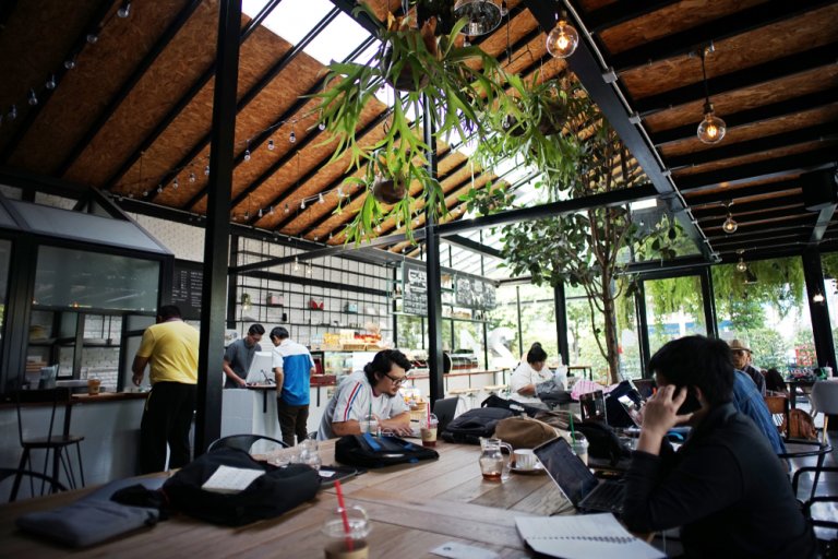 Modern café with a glass roof and wooden beams. Patrons are sitting at tables working on laptops, fostering a coworking environment. A few people are standing at the counter. Hanging plants add greenery while large windows let in natural light. The atmosphere is busy and vibrant.
