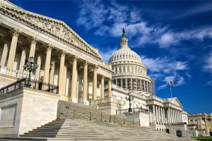 The United States Capitol building in Washington, D.C., with its iconic dome and neoclassical architecture, is set against a clear blue sky. Stairs leading up to the entrance, columns, and the American flag flying on a pole are also visible—much like the structured details found in a federal resume.