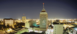 A nighttime cityscape in Fresno County features a tall building with an illuminated "Security Bank" sign at the top. Surrounding the building are various lit buildings, streets, and trees, all against a clear, dark sky.