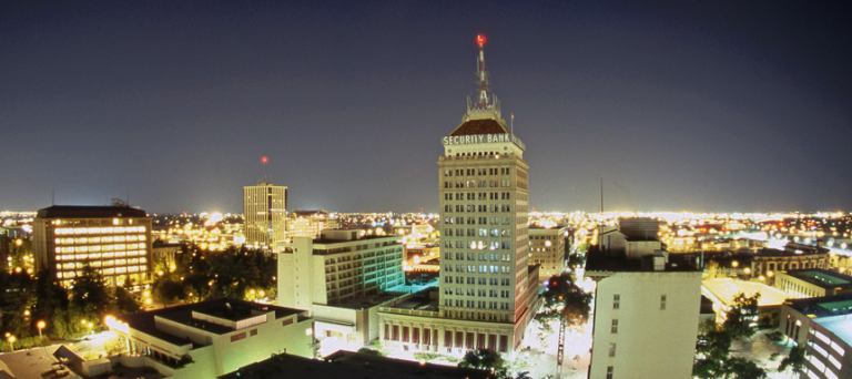 A nighttime cityscape in Fresno County features a tall building with an illuminated "Security Bank" sign at the top. Surrounding the building are various lit buildings, streets, and trees, all against a clear, dark sky.