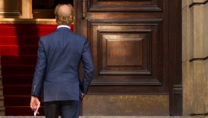 A man with balding hair, dressed in a blue suit, is seen from behind as he walks up a set of stairs towards a large, ornate wooden door. He holds a federal resume in his hand, and a red carpet lines the stairs.