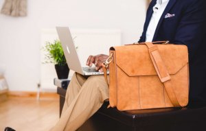 A person in a dark blazer and light-colored pants sits on a bench, typing on a laptop, perhaps working on their federal resume. A tan leather messenger bag rests beside them on the seat. A small green plant is placed on a table next to the person. The setting appears to be indoors.