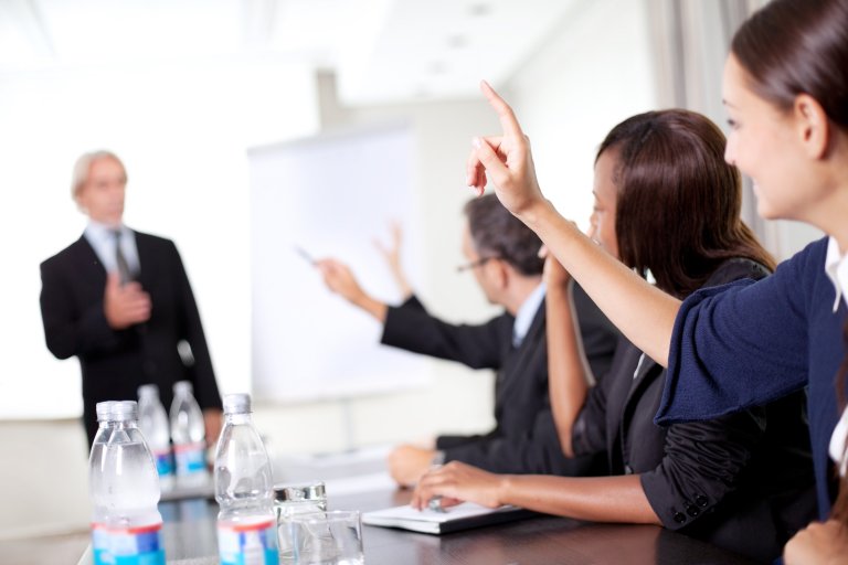 A group of professionals seated at a conference table, some with raised hands, attentively facing a speaker in the foreground. The setting suggests an interactive business meeting or presentation about new jobs programs, with water bottles and notepads on the table.