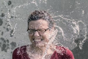 A person wearing glasses and a red shirt is mid-laugh with eyes closed as water splashes over their face, spreading outward dramatically, providing a momentary escape from the stress of their job search. The background is neutral and blurry, focusing attention on the water splash and their joyful expression.