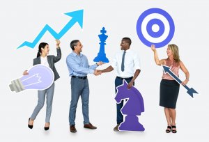 A diverse group of four business professionals stand together holding various large symbols including a light bulb, an upward-trending graph, a chess piece, and a target. Showcasing their soft skills, two men in the center are shaking hands confidently.