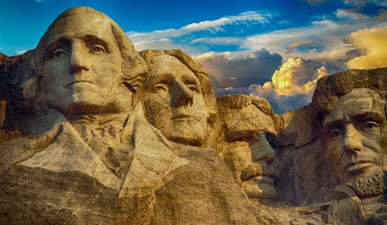 Mount Rushmore National Memorial, featuring the carved faces of four U.S. presidents, stands as a testament to government leadership. The sculptures are embedded in a cliff beneath a partly cloudy sky at sunset, highlighting their detailed expressions.