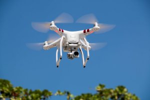 A white quadcopter drone with red stripes is soaring in the sky. Equipped with a camera underneath, its propellers are blurred in motion against a clear blue backdrop, while some greenery frames the bottom scene. This advanced piece of technology could be part of government aerial surveillance efforts.
