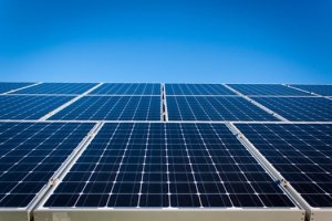 A close-up view of an array of solar panels under a clear blue sky. The panels, supported by government initiatives, are aligned uniformly, reflecting sunlight and capturing renewable energy. The image conveys a sense of clean energy and sustainability.