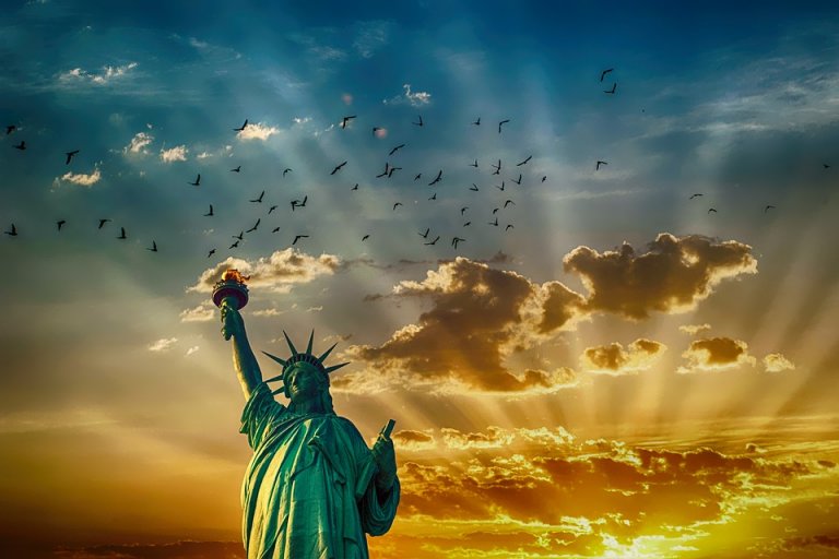 A majestic view of the Statue of Liberty at sunset. The sun sets behind clouds, casting rays of light across the sky. Birds fly above the statue, adding to the dramatic scene. The vivid colors of orange, yellow, and blue enhance the silhouette, reflecting her enduring legacy.
