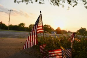Several small American flags are planted in the ground near a sidewalk. The sun sets in the background, casting a warm glow over the scene. Trees and a road are visible in the distance, creating an atmosphere that feels both calm and reflective—a visual legacy of patriotism and tranquility.