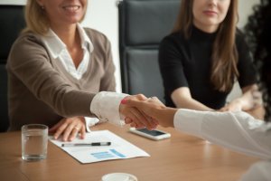 Two individuals are shaking hands across a table during a meeting, with one person partially visible and the other fully shown, accompanied by a third person in the background. A document, pen, and water glass are on the table, indicating a professional setting likely related to human resources.
