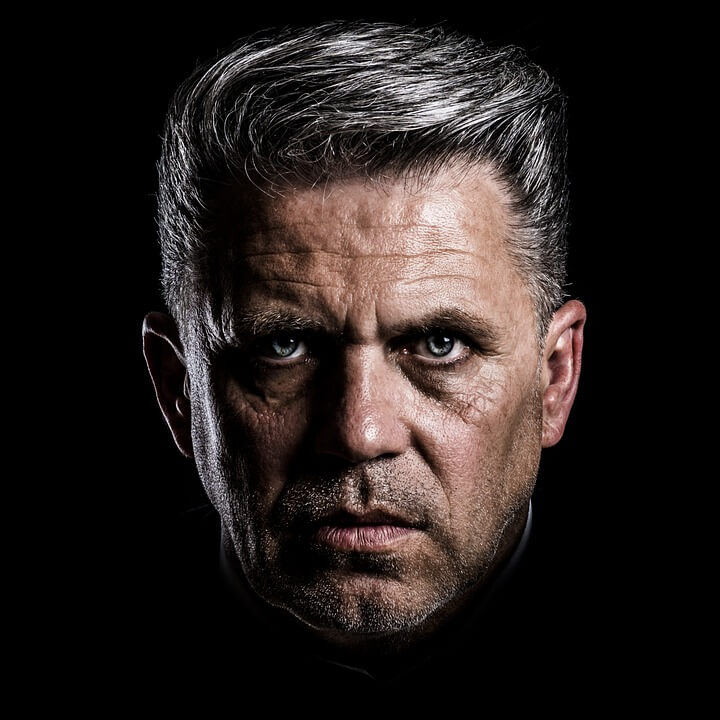 A close-up of a middle-aged man with short, gray hair and intense blue eyes. His face is illuminated against a dark background, highlighting the wrinkles and texture of his skin, giving the image a dramatic and serious tone, evoking the presence of a seasoned leader.