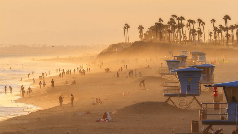 An orange-toned sunset scene of a beach with numerous people walking and relaxing, capturing the essence of endless summer days. Lifeguard towers, symbolizing vigilant employment, are stationed along the sandy shore, with tall palm trees and a hazy horizon in the background. Seagulls soar above the water, enhancing the tranquil atmosphere.