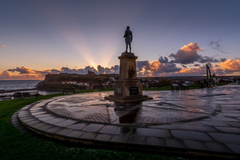 A statue of a captain stands on a paved circular platform overlooking a coastal town at sunset. The sky is filled with colorful clouds, and rays of sunlight break through. In the background, buildings and the coastline are visible, with gentle waves on the sea.