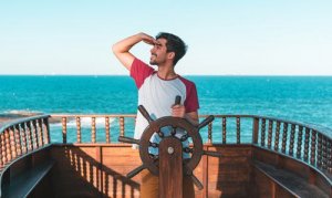 A captain stands at the helm of a wooden ship, gripping the ship's wheel with one hand and shading his eyes with the other as he gazes into the distance. The ocean is visible behind him under a clear, blue sky.