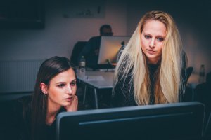 Two women are focused on a computer screen. One, with long blonde hair, is standing and leaning over to look at the screen. The other, with long brown hair, is seated and also gazing intently at the screen. A blurred background shows a human resources office setting.