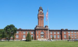 A historic, red-brick building with a tall central clock tower stands proudly against a clear blue sky in Lake County. An American flag flies on a flagpole in front of it. The building is surrounded by a well-maintained lawn with a few trees.