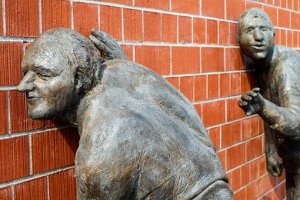 Two bronze statues are positioned against a red brick wall. The statue in the foreground appears to be listening closely with an ear pressed to the wall, while the statue in the background looks as if it is stealthily approaching, perhaps eager to listen in on a clandestine conversation.