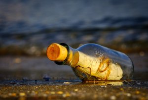 A glass bottle with a cork, containing a rolled parchment inside, lies on wet sand at the beach with gentle waves in the background. The bottle is partially covered with droplets and reflecting the light, as if waiting for someone to listen to its timeless message.