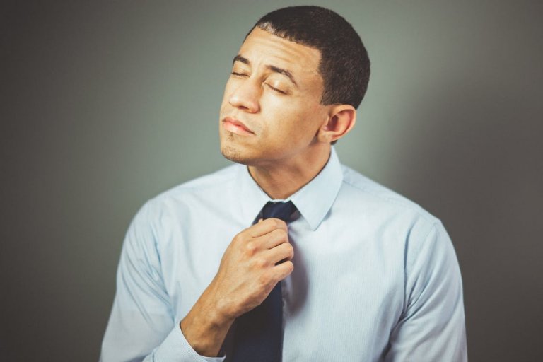 A man with short dark hair, wearing a light blue dress shirt and a dark tie, is adjusting his tie with one hand. His eyes are closed, and he has a contemplative expression on his face, perhaps reflecting on upcoming meetings. The background is plain and gray.