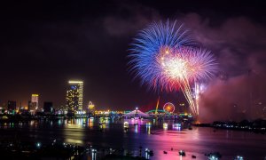 A vibrant nighttime cityscape featuring a river with boats, high-rise buildings with illuminated windows, a lit-up bridge, and a glowing Ferris wheel. Amid this stunning display of colorful fireworks lighting up the sky, offices of major human resources firms dot the skyline.