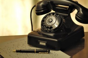 A vintage black rotary dial telephone sits on a wooden surface beside a handwritten letter and a fountain pen. The telephone, almost expressive in its stillness, features a circular dial with numbers and a coiled cord connecting the handset to the base, evoking an era when body language spoke volumes.
