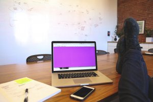 A person relaxes with their legs up on a wooden conference table, next to a laptop with a purple screen. An open notebook, smartphone, and pen are scattered on the table. The whiteboard in the background is covered in handwritten notes and flowcharts, subtly conveying confident body language.