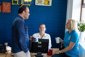 Three people interacting in an office, with a blue wall and a window with plants in the background. One man and one woman are standing, holding mugs, while another woman sits at a desk with a computer, smiling. Their body language indicates a friendly and casual atmosphere.