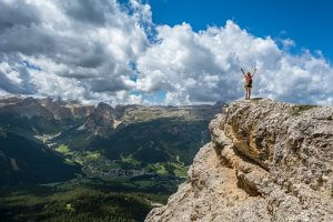 A person stands triumphantly with arms raised at the edge of a rocky cliff, overlooking a vast, mountainous landscape under a partly cloudy sky. Green valleys and distant peaks stretch out below, highlighting the expansive natural beauty—a metaphor for climbing the peaks of one's career.