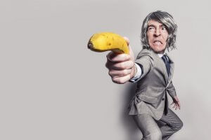 A person with gray hair is dressed in a light gray suit and white dress shirt, adopting a dramatic stance as they extend a banana in their hand as if pointing it like a gun, against a plain white background. Their intense and serious expression suggests they're ready for any career obstacle.