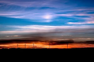 A stunning sunset with a brilliant mix of orange, pink, and blue hues in the sky. Silhouetted against the colorful backdrop are several wind turbines lined up on the horizon, highlighting the contrast between nature and renewed energy technology.