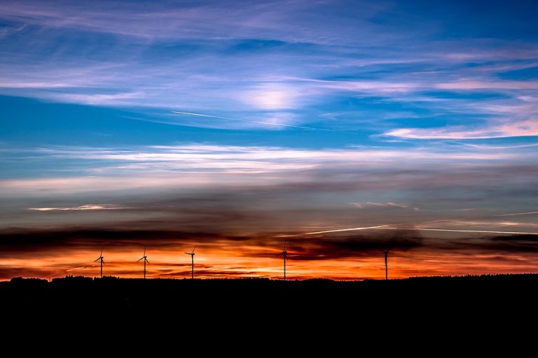 A stunning sunset with a brilliant mix of orange, pink, and blue hues in the sky. Silhouetted against the colorful backdrop are several wind turbines lined up on the horizon, highlighting the contrast between nature and renewed energy technology.