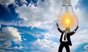 A silhouette of a person in a suit stands on a grassy field, appearing to hold up a large, renewed light bulb that is illuminated against a backdrop of a bright blue sky with scattered clouds. The image represents creativity and innovation.