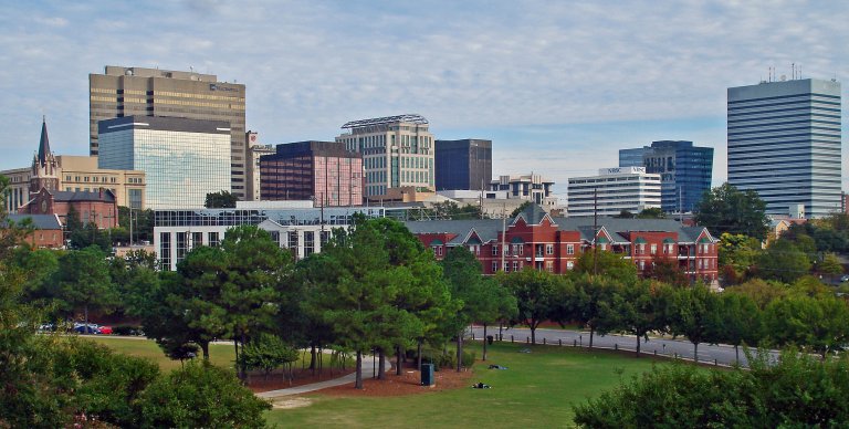 A cityscape featuring modern high-rise buildings mixed with older architectural structures. In the foreground, there is a lush green park with trees and a pathway, reminiscent of the serene landscapes found in Lake County. The skyline is under a partly cloudy sky.