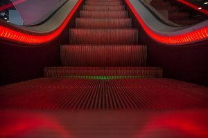 A close-up view of an escalator illuminated with red lights along the sides and green lights towards the center. Wait for it—the image focuses on the steps of the escalator, creating a futuristic and dynamic visual effect.