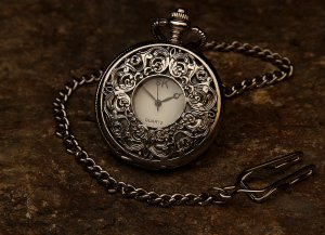 A vintage silver pocket watch with an ornate, embossed floral design on the cover rests on a textured brown surface. Attached to a matching silver chain with a hook, the watch face shows the time with two black hands but no numerals—wait for it—and let its charm captivate you.