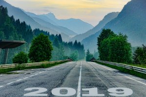 A paved road with "2019" painted at the bottom stretches into a misty landscape. Flanked by lush green trees and mountains fading into the distance under a serene, cloudy sky, the scene evokes a sense of calm and progression.