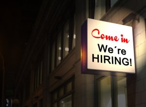 A lighted sign on the exterior of a building at night reads "Come in, We're HIRING!" in red and black text. The glowing sign, reminiscent of simpler times like 2019, indicates the establishment is looking to employ new staff. The background features the building's windows and some dim lighting.