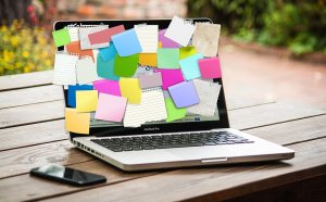 A 2019 laptop is open on a wooden desk, its screen covered with numerous colorful sticky notes and pieces of paper, obscuring any visible content. A smartphone is placed next to the laptop, and the background shows a blurred outdoor scene.