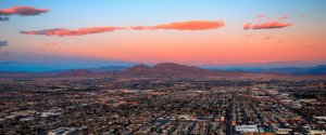 Aerial view of a vast urban landscape in Clark County at dusk, with a painted sky in shades of orange, pink, and blue. Scattered clouds glow in the sunset, and mountains form a scenic backdrop behind the sprawling city.