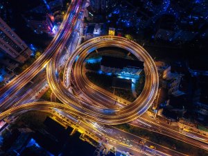 Aerial view of a city at night featuring a prominent circular highway interchange illuminated by streetlights. Various roads chaos the interchange with streaks of vehicle lights highlighting the bustling traffic. Buildings and city lights are visible in the surrounding area.