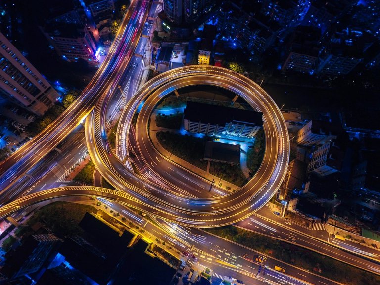 Aerial view of a city at night featuring a prominent circular highway interchange illuminated by streetlights. Various roads chaos the interchange with streaks of vehicle lights highlighting the bustling traffic. Buildings and city lights are visible in the surrounding area.