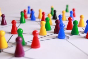 Colorful game pieces lie scattered in a seemingly chaotic arrangement on a board with interconnected lines and nodes, representing a network or connected system. The vibrant pieces—red, blue, green, yellow, purple, and black—create an intricate tapestry of organized chaos.