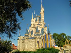 A majestic castle with blue and gold turrets stands against a clear blue sky. Surrounded by lush trees and greenery, flags with medieval designs decorate the front. The architecture features detailed stonework and spires, creating a fairy-tale atmosphere that sparks employee engagement through its enchanting allure.