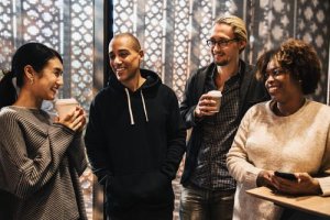 Four people are standing together, smiling and talking. One person is holding a clipboard and another is holding a phone. Two others are holding coffee cups. They seem to be having a friendly conversation in a relaxed environment with patterned walls in the background, fostering strong employee engagement.