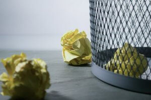 A wire mesh trash can with crumpled yellow paper balls and scattered files inside and around it on a gray wooden floor. The background is blurry, focusing attention on the trash can and the discarded items.