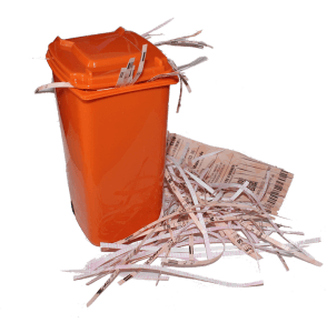 An orange waste bin with a partially open lid is overflowing with shredded paper. Strips of shredded paper and fragments of files are scattered outside the bin, indicating it is full. Nearby, some larger pieces of paper with text are also visible.
