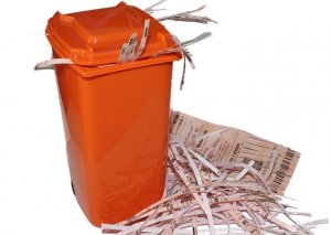 An orange trash bin filled with shredded paper pieces and scattered files is spilling out onto the floor. The bits of documents and printed material are strewn around the bin, set against a white background.