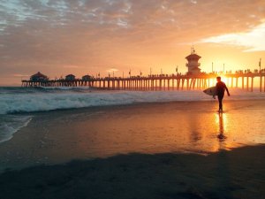 A surfer carrying a surfboard walks along Huntington Beach towards the ocean at sunset. In the background, a large pier extends into the water, populated with numerous people. The sky is painted with hues of orange and gold as the sun begins to set.