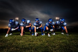 Five American football players wearing blue jerseys and helmets are positioned in a line of scrimmage on a grassy field under a dark, cloudy sky. The middle player is preparing to snap the football while the kicker readies himself nearby. Stadium lights glow in the background.